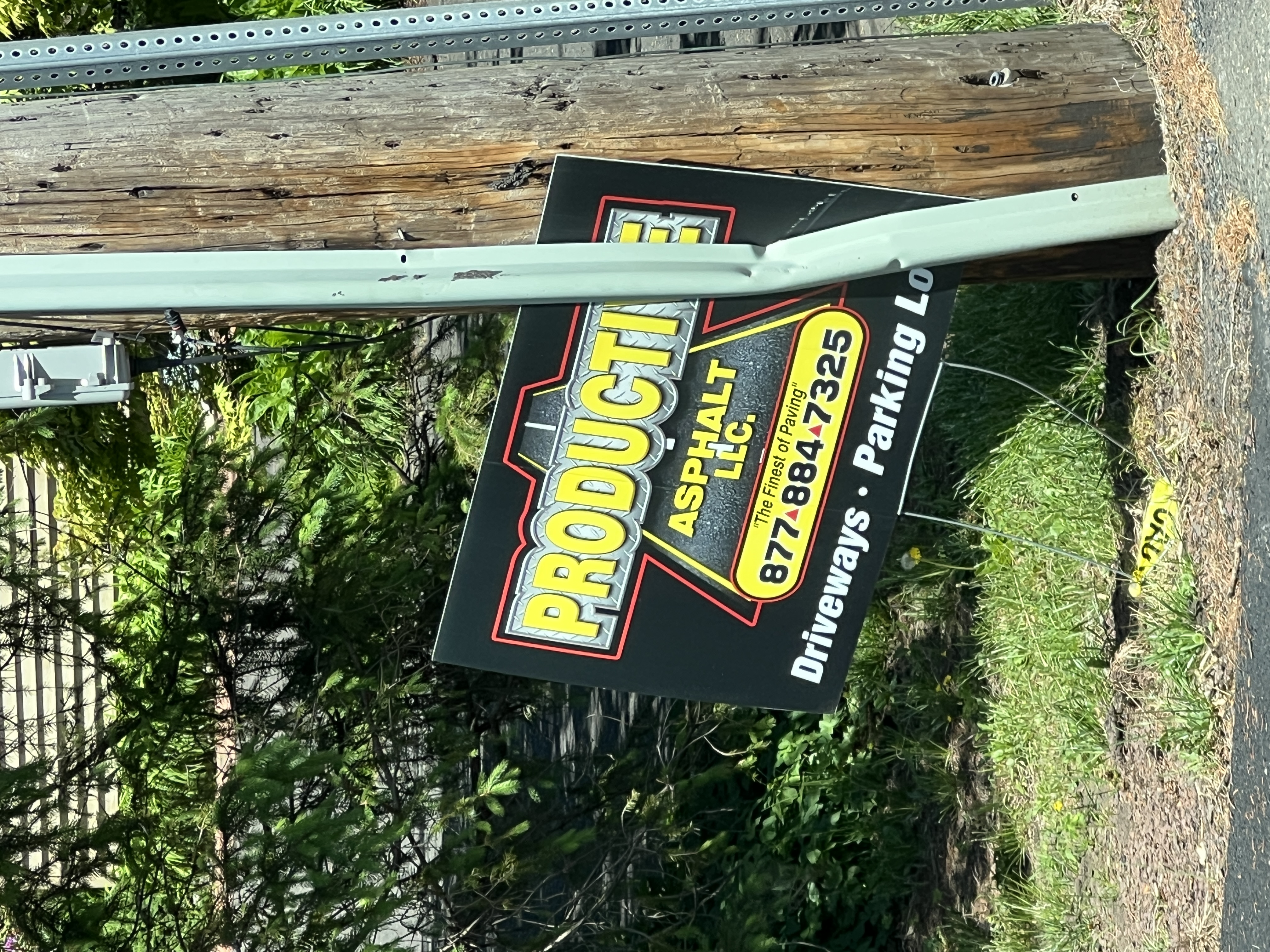 Yard sign for Productive Asphalt LLC nailed to a telephone pole — black background with diamond-plate wordmark, tagline 'The Finest of Paving', 877-884-7325, driveways and parking lots.