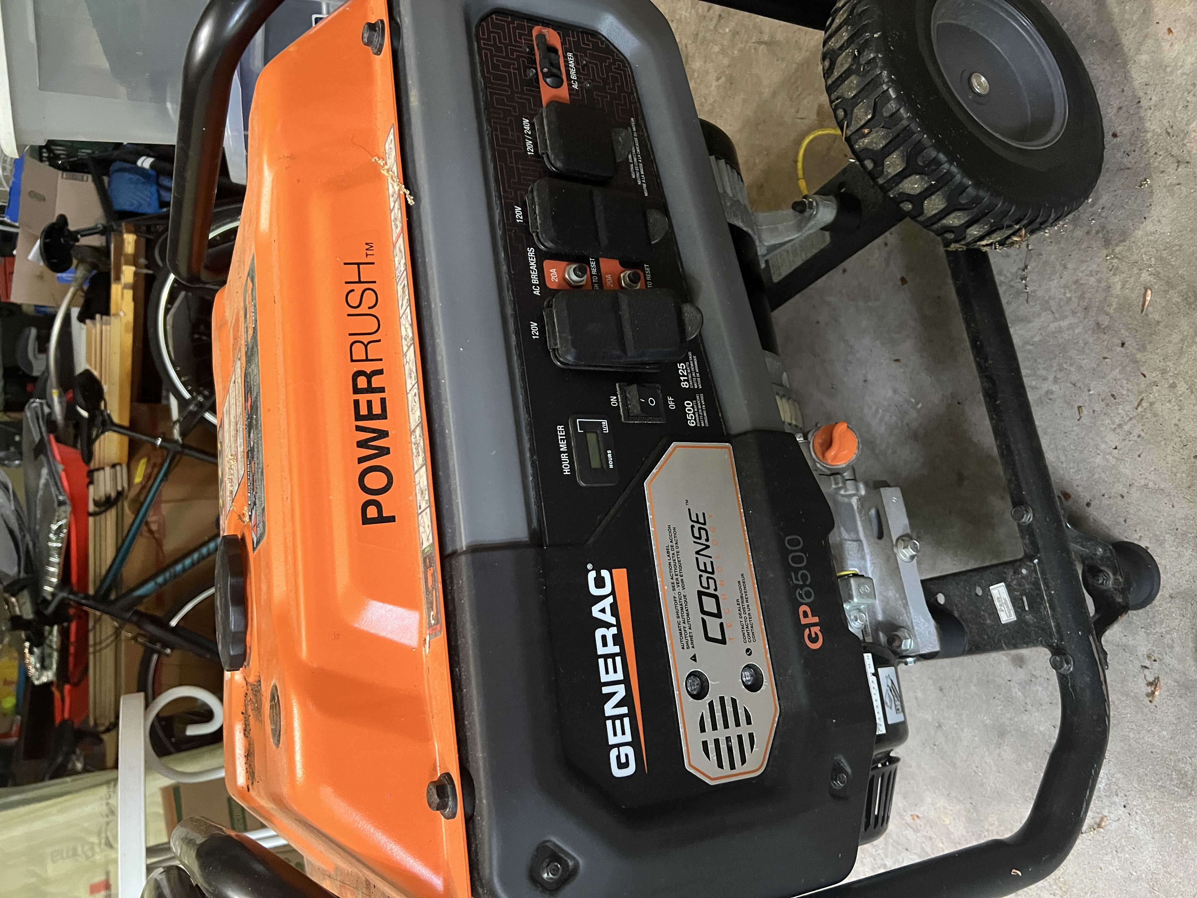 Generac GP6500 portable gas generator on its wheeled frame in the New Hope garage. Orange POWERRUSH housing with control panel showing 120V/240V outlets, AC breakers, hour meter, and the COsense automatic-shutoff badge.